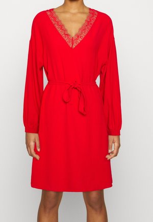 Kvinde iført en rød langærmet kjole med blonde-V-halsudskæring og bundet taljebælte, stående mod en ensfarvet lys baggrund.