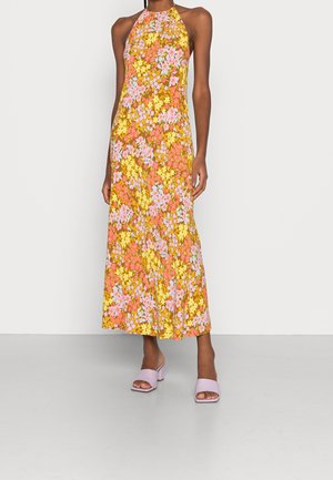 Femme portant une robe midi sans manches à col halter avec des motifs floraux roses, jaunes et orange, associée à des talons ouverts violet clair.