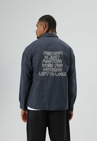 Overhemd met knopen in een donkerblauw en zwart geruit patroon, met witte geborduurde tekst op de achterkant. Lange mouwen, relaxte pasvorm.