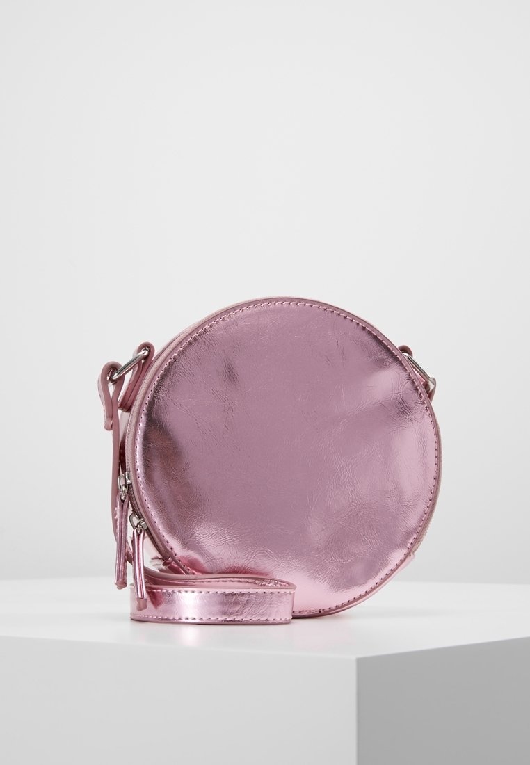 Bolso redondo en material metálico rosa, con una textura suave, cierre de cremallera y una correa desmontable. Diseño minimalista sin hardware visible.