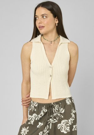Femme portant un haut sans manches crème côtelé avec boutons et un pantalon ample olive avec des motifs floraux blancs, regardant sur le côté avec la main dans le dos.