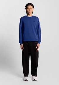 Blauwe sweatshirt met een ronde halslijn en zwart logo op de borst, gecombineerd met zwarte broeken en lichtgekleurde sneakers.