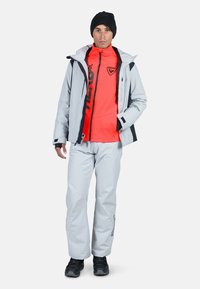 Herrars utomhusoutfit bestående av en ljusgrå jacka, ett klart orange innerlager, grå byxor och svarta skor. Jackan har huva och dragkedjor.