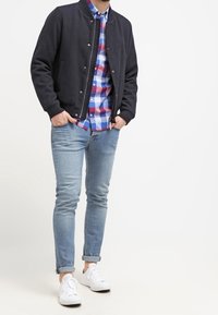 Marineblauer Bomberjacke, kariertes Hemd in Blau und Rot, hellblaue Jeans und weiße Turnschuhe. Lässiges Schichten und figurbetonte Silhouette.