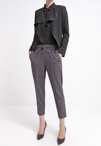 Chaqueta corta negra con frente drapeado, combinada con pantalones gris ajustados con cintura con cordón. Tacones altos negros completan el atuendo.