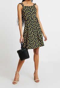 Svart klänning med gult blommönster, ärmlös design, rynkade axelband, utsvängd kjol, matchad med en svart handväska och nude-hälar.