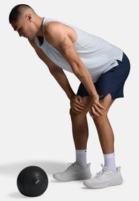 Mand iført hvid tanktop og marineblå shorts bøjer sig fremad med hænderne på knæene, hvilende ved siden af en sort træningsbold på gulvet.