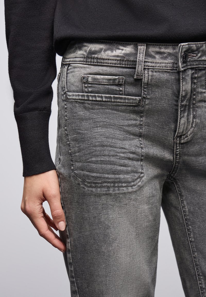 Jeans en denim gris avec une texture délavée, présentant des détails sur les poches arrière et un design slim. Portés avec un pull noir.