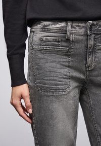 Jean gris de denim con una textura desgastada, que presenta detalles en los bolsillos traseros y un diseño de corte ajustado. Llevado con un suéter negro.