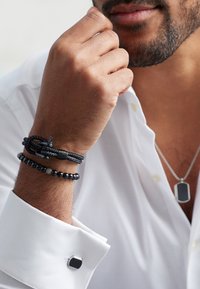 Black bracelets made of beads and braided cord on a wrist, paired with a silver chain and rectangular pendant. White shirt cuff with cufflink.