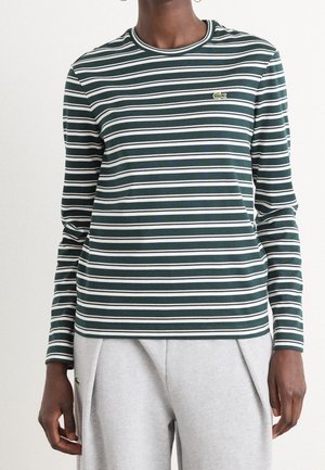 Personne portant un haut à manches longues rayé vert foncé et blanc et un pantalon gris clair avec un petit logo sur les deux vêtements.