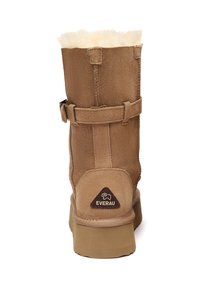 Tan suede boot with a cream faux fur lining, a side strap, and a rubber sole. Features a brown triangular logo on the back.