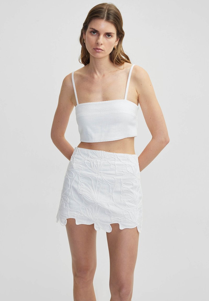 Top blanco corto con tirantes finos y falda mini blanca bordada con patrones florales y dobladillo en forma de ondas. Materiales ligeros y texturizados.