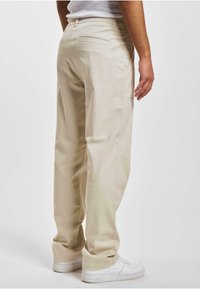 Beige-housut rentoa tyyliä, valmistettu sileästä kankaasta. Takana taskut ja lahkeet lepäävät valkoisten sneakersien päällä. Yksinkertainen ja funktionaalinen muotoilu.