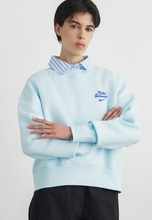 Tânăr cu păr scurt și închis la culoare, purtând un hanorac Nike Summer de culoare albastru deschis peste o cămașă cu dungi, cu brațele încrucișate și o expresie neutră.