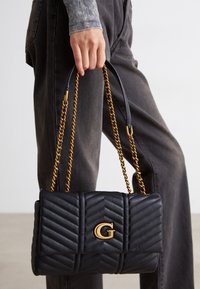 Hånd der holder en sort quiltet skuldertaske med guld kædestrop og guld "G" logo, båret med mørkegrå bukser og en stribet bluse med lange ærmer.