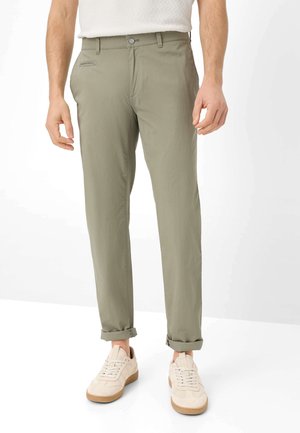 Man draagt opgerolde lichtolijfgroene chino's en beige sneakers, staand tegen een effen witte achtergrond.