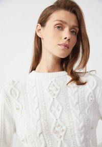 Hvit strikket genser med kabelstrikkdesign, perlepynt og klassisk rund hals. Glatt tekstur med intrikate mønster.