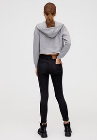 Sweat à capuche gris coupé, avec une capuche, associé à un jean skinny noir avec des poches arrière. Étiquette blanche visible à la taille, porté avec des baskets noires.