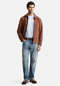 Rjava rebrasta jakna čez svetlo modro majico, kombinirana s svetlo modrimi jeans hlačami in rjavimi supergami z belim vzorcem.