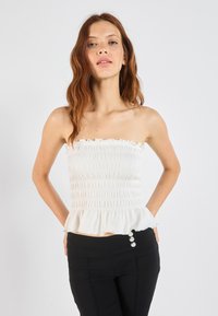 Rihmata ilma varrukat valge topp, millel on volditud tekstuur, laineline kaelus ja peplum alläär. Kombineeritud mustade pükstega, millel on kolm dekoratiivset nööpi.