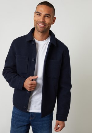 Smilende mand iført en marineblå jakke med knapper over en hvid T-shirt og blå jeans mod en ensfarvet lys baggrund.