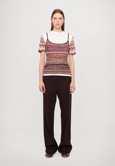 Top multicolor cu model în zigzag. Guler alb, pantaloni relaxați cu model maroniu și croială largă. Modelul stă pe un fundal simplu.