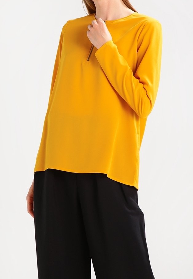 Světle žlutý top s dlouhými rukávy, kulatým výstřihem a jemnými švy, doplněný volnými černými kalhotami. Viditelná hladká textura látky.