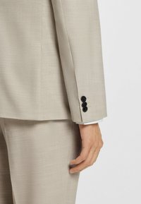 Lys beige blazer med en tekstureret overflade. Har en manchet med tre sorte knapper og en hvid skjortemanchet, der titter frem nedenunder.