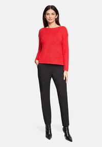 Frau steht mit den Händen in den Taschen, trägt einen roten Strickpullover, schwarze Hose und schwarze Stiefeletten vor weißem Hintergrund.