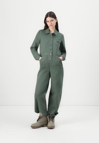 Combinaison verte avec col, boutons à l'avant et deux poches poitrine ; associée à des bottines beiges à lacets, avec une semelle robuste.