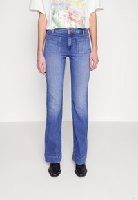 Jean flare bleu clair avec deux poches arrière, taille mi-haute et texture légèrement délavée. Associé à des bottes noires à bout pointu.