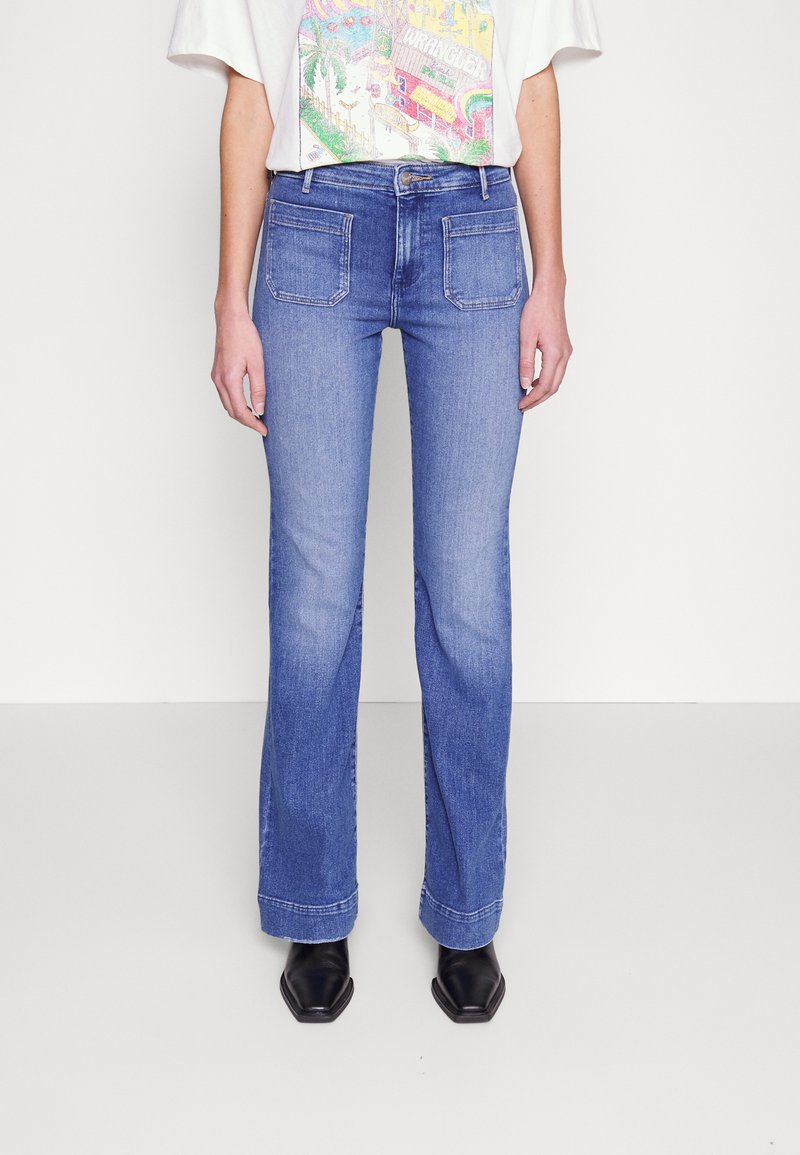 Jean flare bleu clair avec deux poches arrière, taille mi-haute et texture légèrement délavée. Associé à des bottes noires à bout pointu.