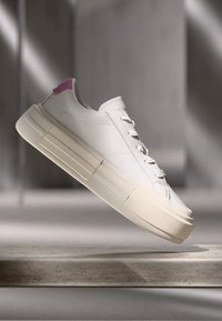 White low-top sneaker with cream sole and pink heel detail, shown floating above a stone surface in soft lighting.
