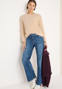 Jasnobeżowa koszula z długim rękawem, niebieskie dzwoniaste jeansy i białe trampki z czerwonymi akcentami. Model trzyma ciemnofioletowy sweter. Gładkie tekstury.