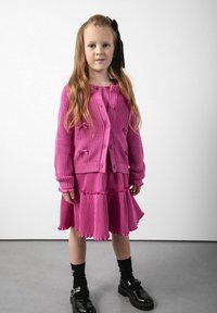 Jasnoróżowy sweter z dzianiny z okrągłymi guzikami, zestawiony z plisowaną różową sukienką warstwową, czarnymi skarpetkami i błyszczącymi czarnymi butami.