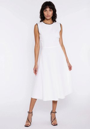 Kobieta o ciemnych, kręconych włosach, ubrana w bezrękawową białą sukienkę midi z czarną lamówką oraz czarne, rzemykowe szpilki, stojąca na białym tle.