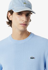 Lichtblauwe trui met geribde kraag en klein geborduurd Lacoste-logo. Draagt een vervaagde blauwe denim pet met hetzelfde logo.