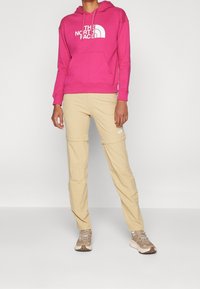 Sudadera con capucha de algodón rosa con logo blanco, combinada con pantalones beige con cremallera para convertirlos en pantalones cortos. Pies calzados con zapatillas beige y marrones. Elementos de diseño minimalista.