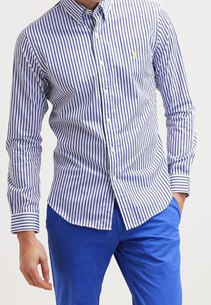 Muž nosící přiléhavou dlouhosrstou modro-bílou pruhovanou košili na knoflíky a modré kalhoty, s jednou rukou v kapse.