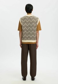 Veste tricotée à motif chevron en beige et marron, portée sur un T-shirt marron, associée à un pantalon marron foncé et des chaussures brillantes.