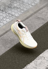 White athletic shoe with pink accents, textured upper, and a slim silhouette. Features laces and a light yellow sole with orange detailing.