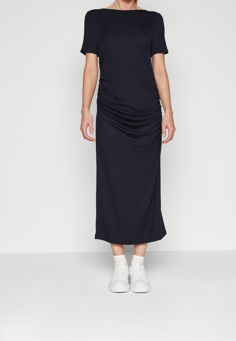 Femme portant une robe midi noire à manches courtes froncée, des chaussettes blanches et des baskets blanches, debout devant un fond clair uni.