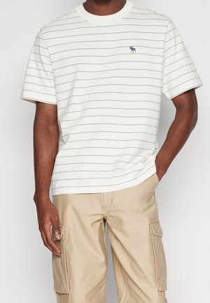 Wit T-shirt met strepen en een ronde hals, voorzien van lichtgrijze strepen en een klein blauw logo op de borst. Gecombineerd met kaki cargobroek.