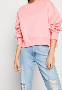 Sudadera rosa recortada con cuello redondo y puños acanalados. Presenta un pequeño logo blanco de Nike en el pecho. Se lleva con vaqueros azules claros rotos.