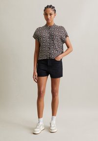 Bloemige blouse met korte mouwen op een zwarte achtergrond, gecombineerd met zwarte denim shorts en witte sneakers. Heeft een knoopsluiting aan de voorkant en een relaxte pasvorm.