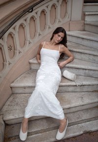 Robe diaphane à broderies florales blanches avec des bretelles fines, accompagnée de ballerines blanches et d'un petit sac à main, assise sur des marches en pierre.
