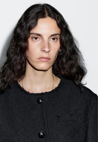 Mladá osoba s dlouhými kadeřavými tmavými vlasy, oblečená v černé texturované sako na knoflíky a zlatými náhrdelníky, na jednoduchém bílém pozadí.
