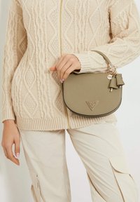 Borsa a tracolla verde oliva con finitura texturizzata, accessori in oro e dettagli con nappine, tenuta da una persona che indossa un maglione a trecce color crema.