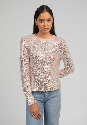 Femme aux longs cheveux bruns portant un haut à manches longues rose pâle orné de sequins et un jean bleu clair, debout devant un fond gris uni.